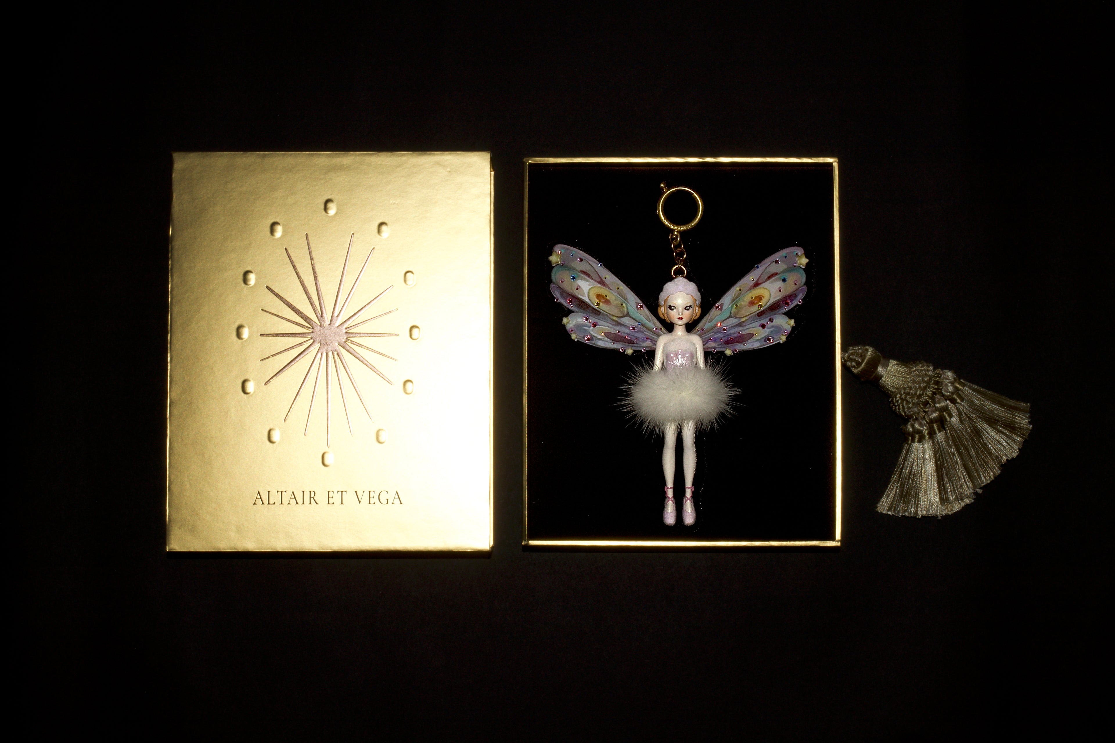 A decorative ballet fairy keychain displayed beside its gold-toned box and a matching tassel on a black background. The keychain features a small doll with pastel wings, a fluffy white tutu, and a gold ring at the top.