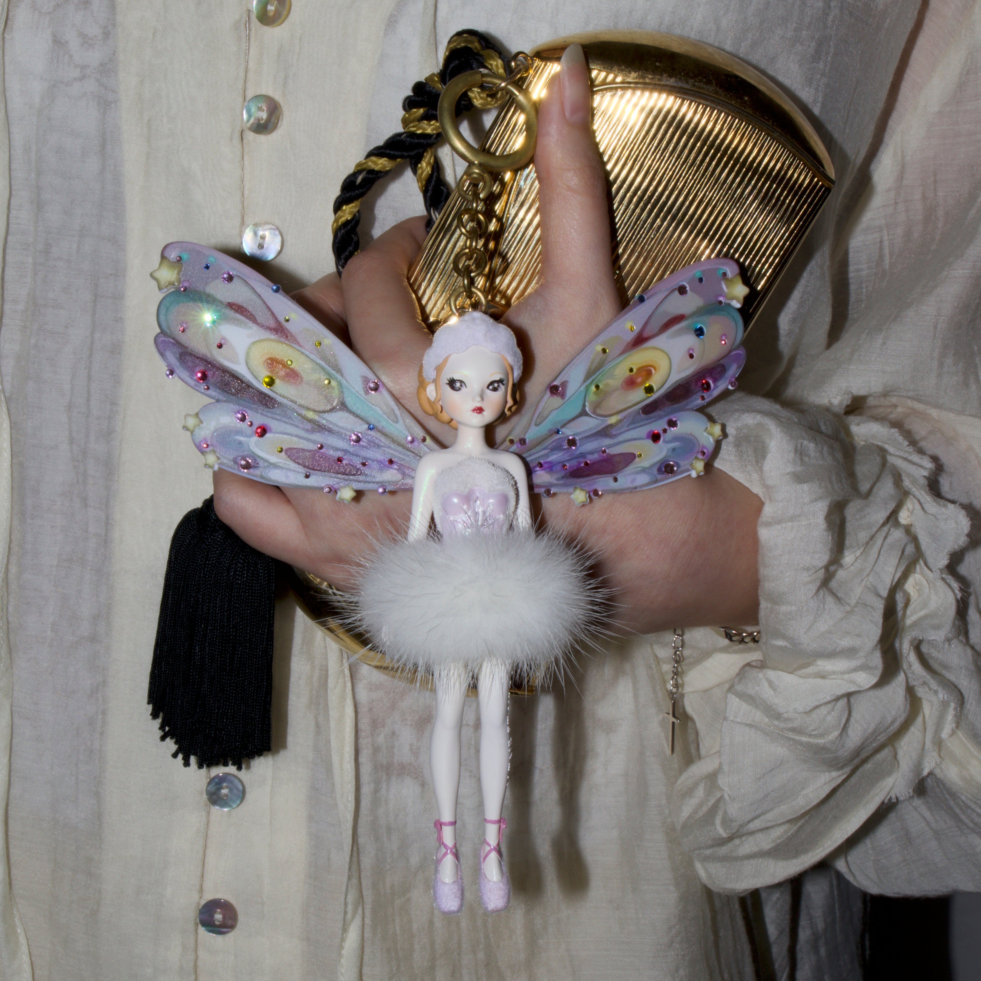 A person in a cream-colored button-up shirt holds a whimsical fairy doll keychain or bag charm. The charm features pastel iridescent wings with rainbow patterns, a white fluffy skirt, and a small doll figure, alongside a gold metallic clutch bag with a black ribbon.