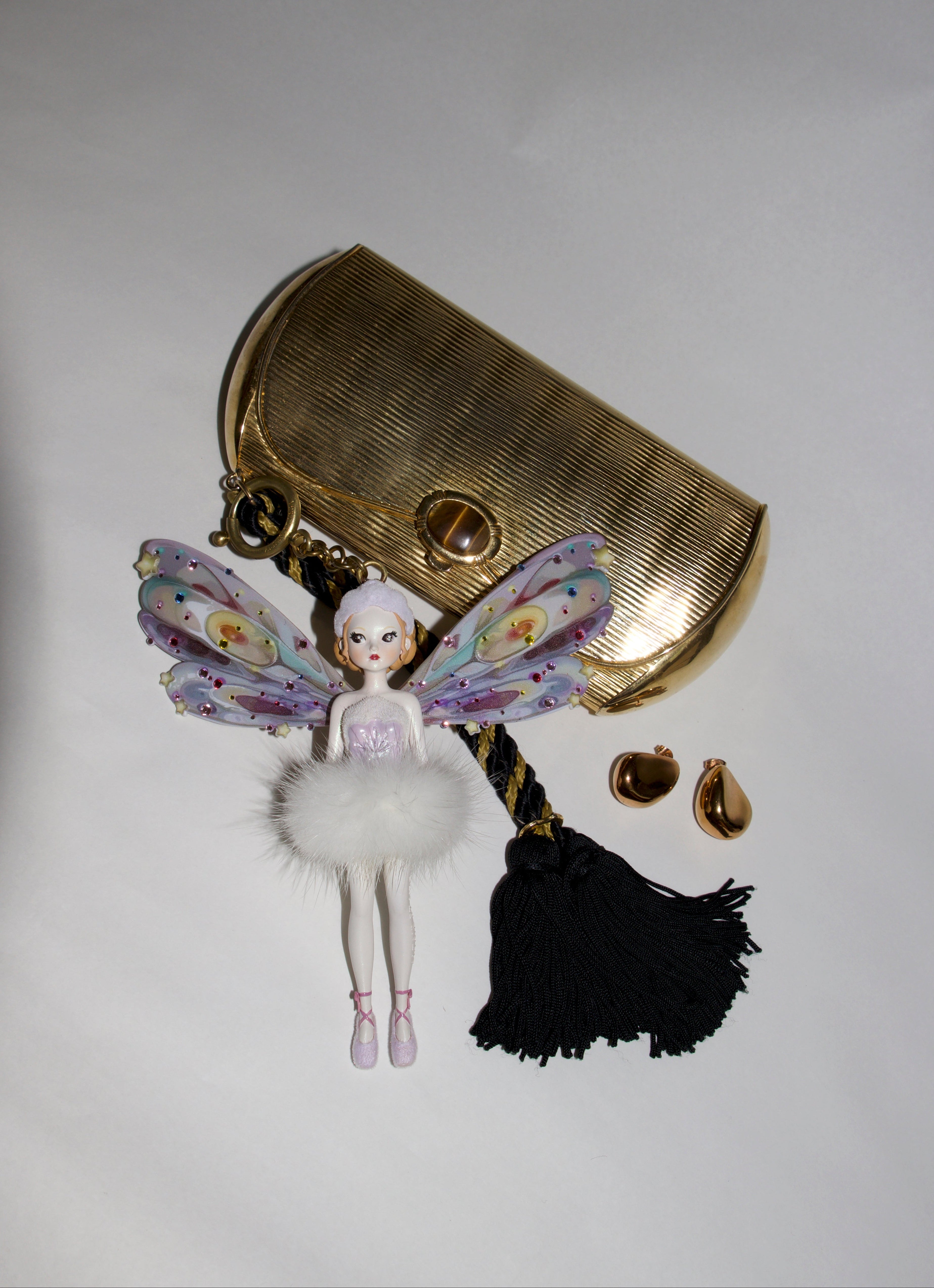 A gold-toned metal clutch bag with a hanging fairy charm, black tassel, and two small gold teardrop earrings, arranged on a plain light background.